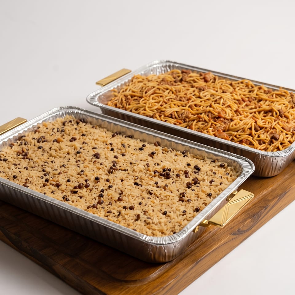 Two catering trays, one with rice and black beans, the other with spaghetti, on a wooden board.