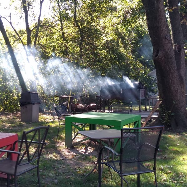 Outdoor dining area with green and red tables, chairs, and smoke from a grill under sun-dappled trees.