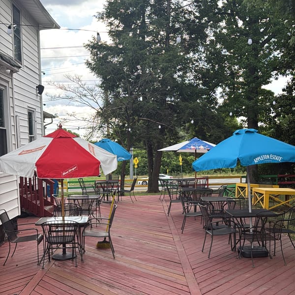 Outdoor patio with red wooden deck, black metal tables and chairs, and colorful umbrellas under trees.
